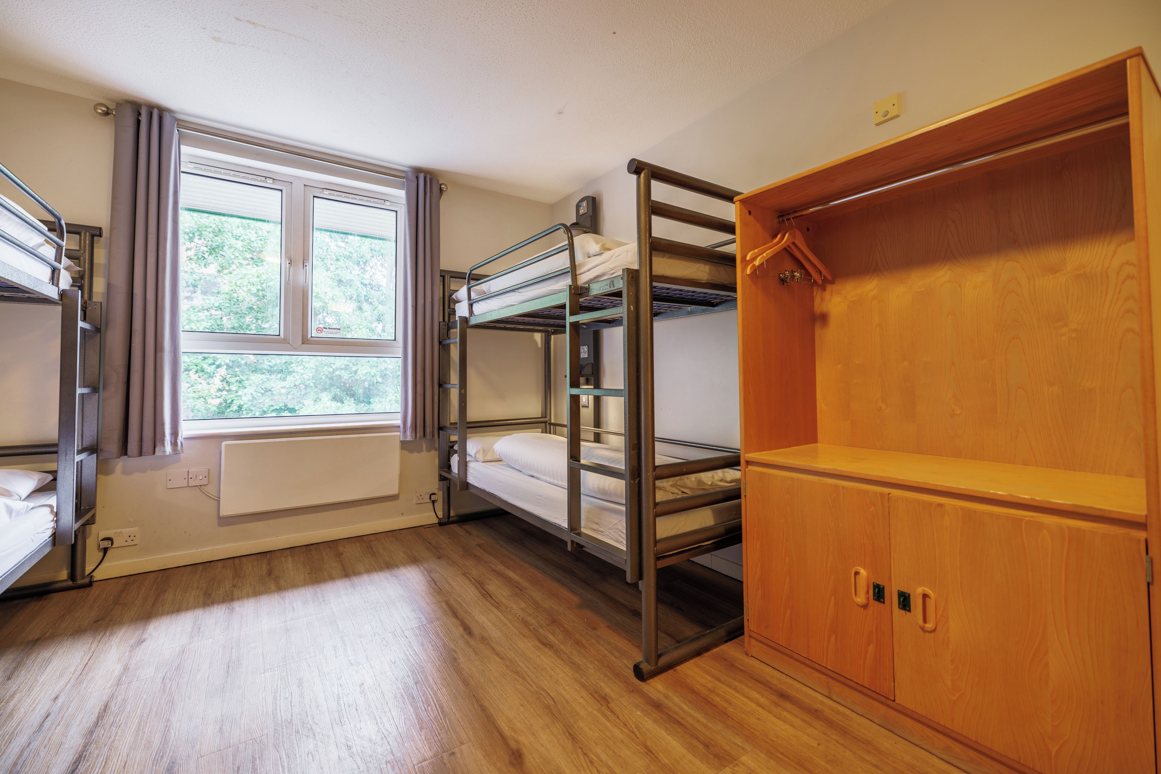 A dormitory room with bunk beds, a radiator, a window with curtains, and a wooden wardrobe.