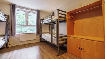 A dormitory room with bunk beds, a radiator, a window with curtains, and a wooden wardrobe.