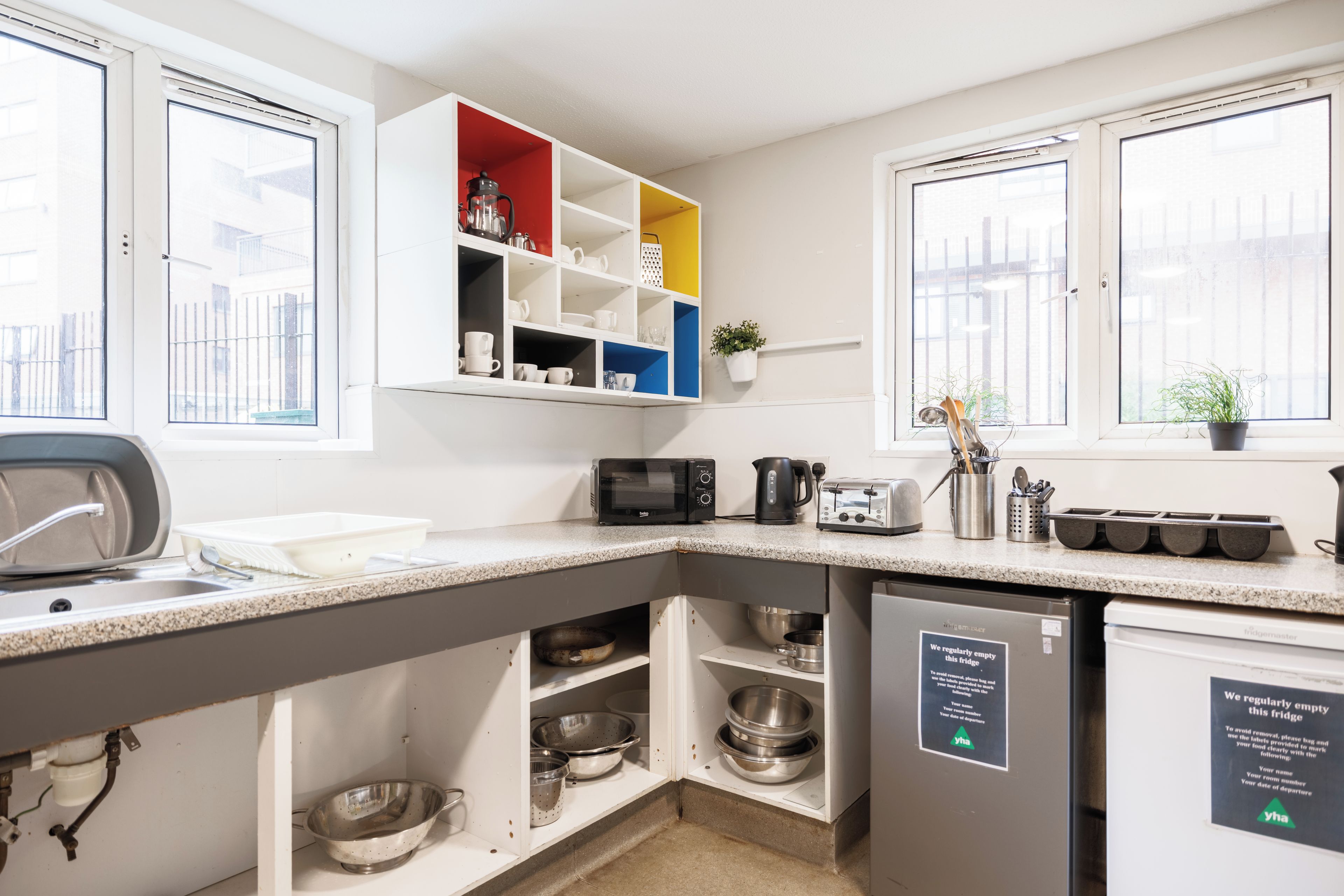 A kitchen with a built-in microwave, toaster, and kettle, a sink, and a fridge with a sign indicating it is regularly emptied.