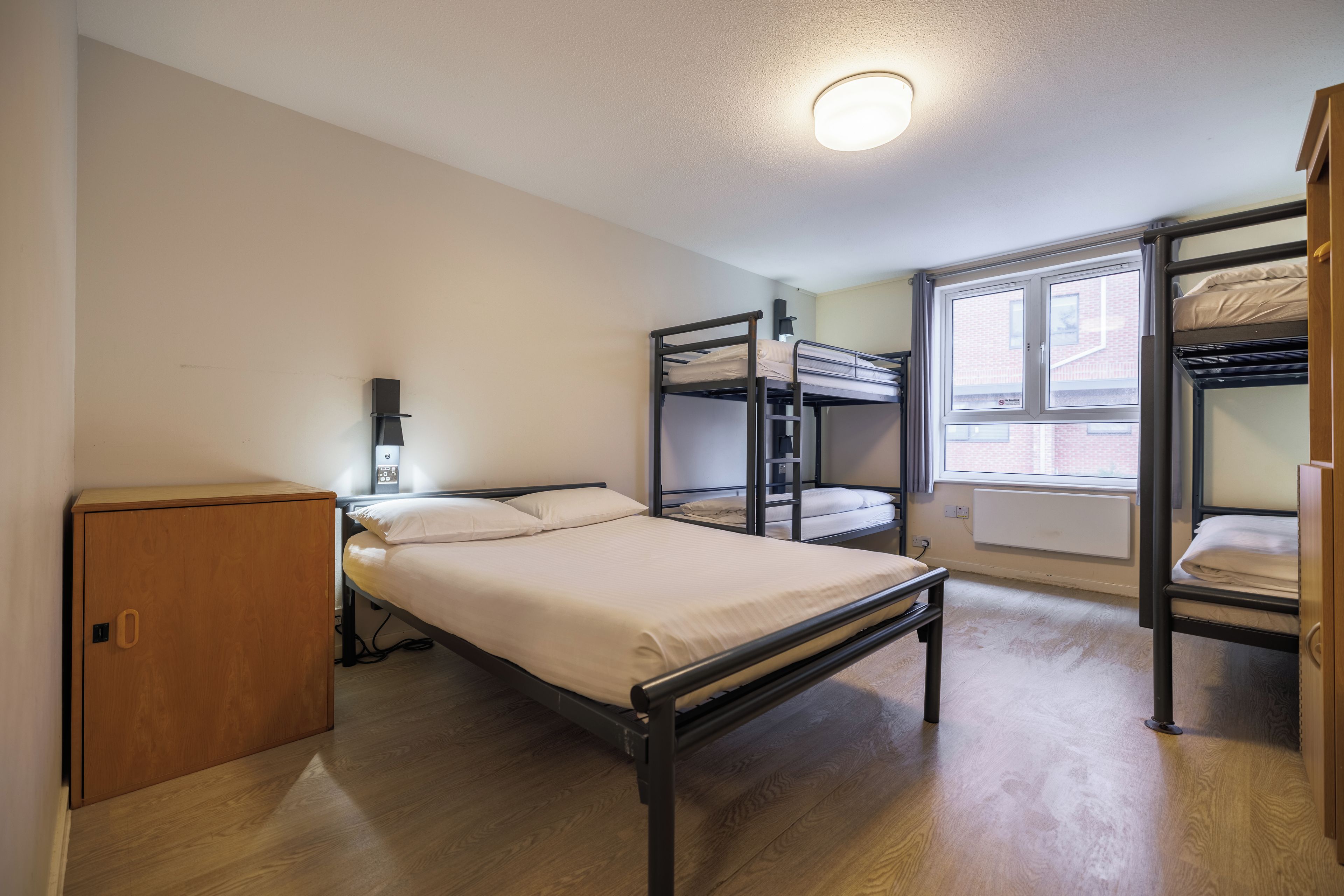 A dormitory room with bunk beds, a single bed, a wooden cabinet, a wall-mounted heater, and a window with a view of buildings.