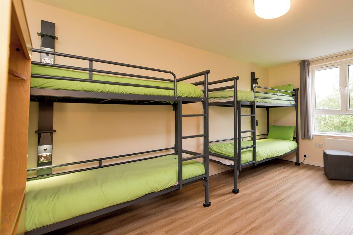 A dormitory room with bunk beds, a wooden floor, a window with curtains, and a ceiling light.