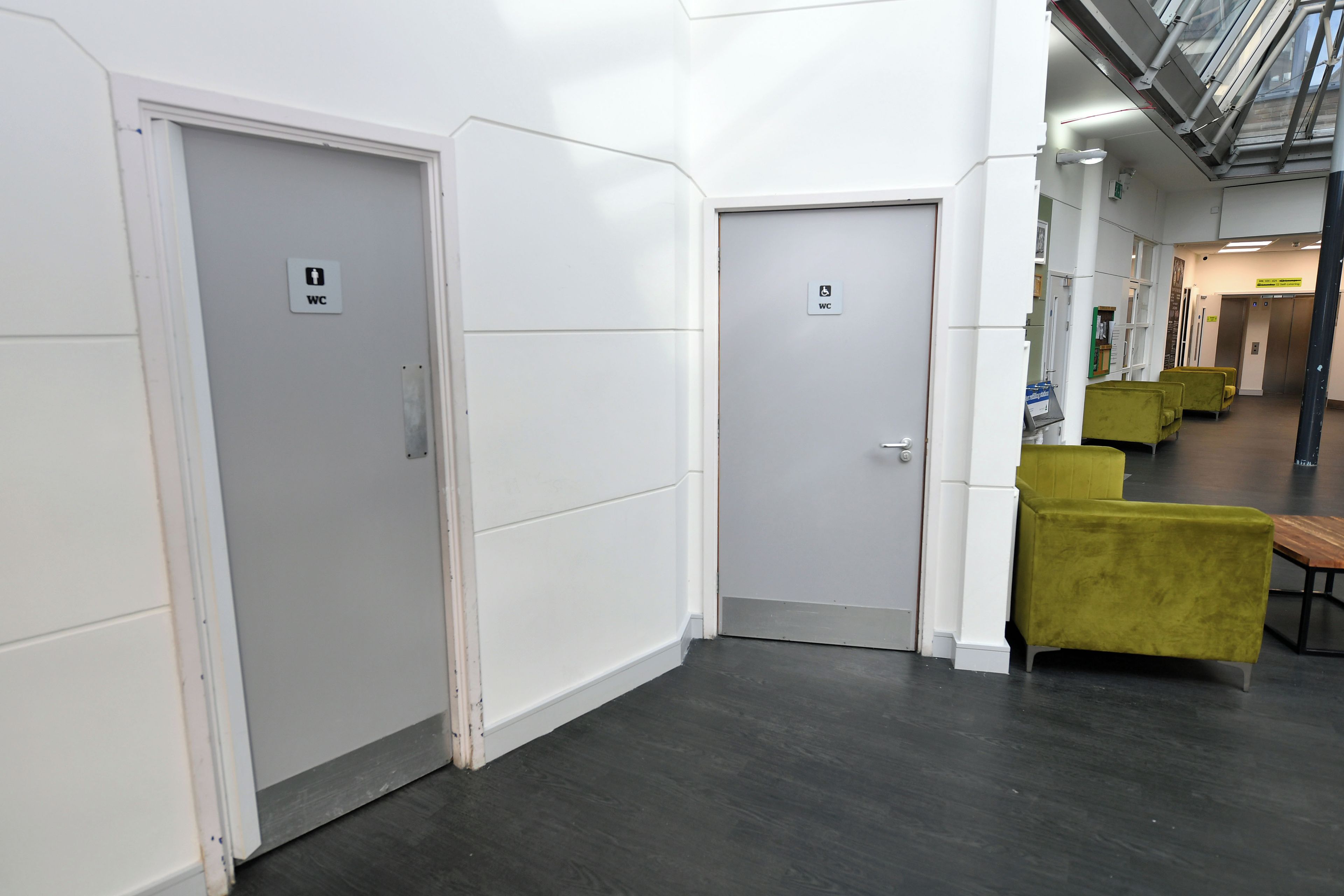 A hallway with two doors, one marked with a WC sign, and a seating area with green chairs.