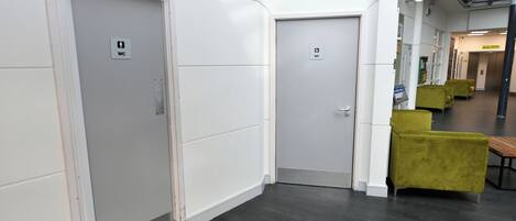 A hallway with two doors, one marked with a WC sign, and a seating area with green chairs.