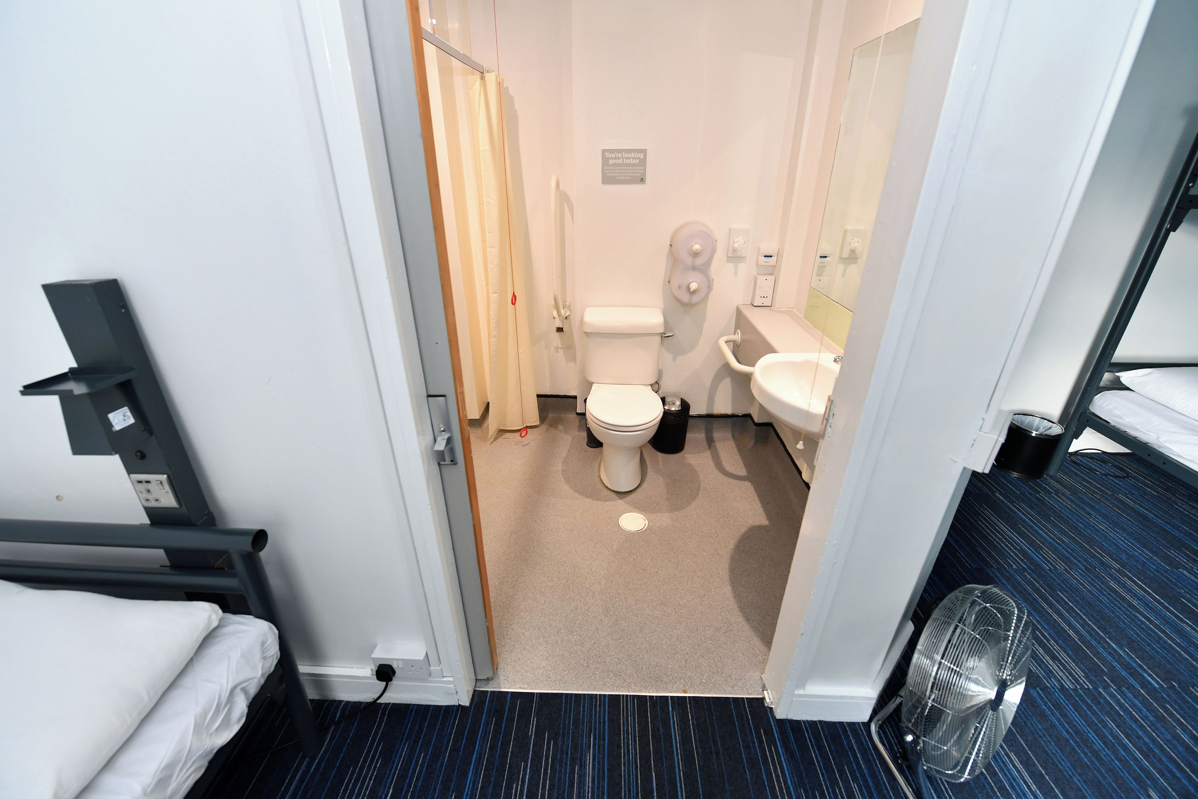 Casa de banho de hotel com sanita, bidé e lavatório. Tem cortina de chuveiro, porta-toalhas e um ventilador.
