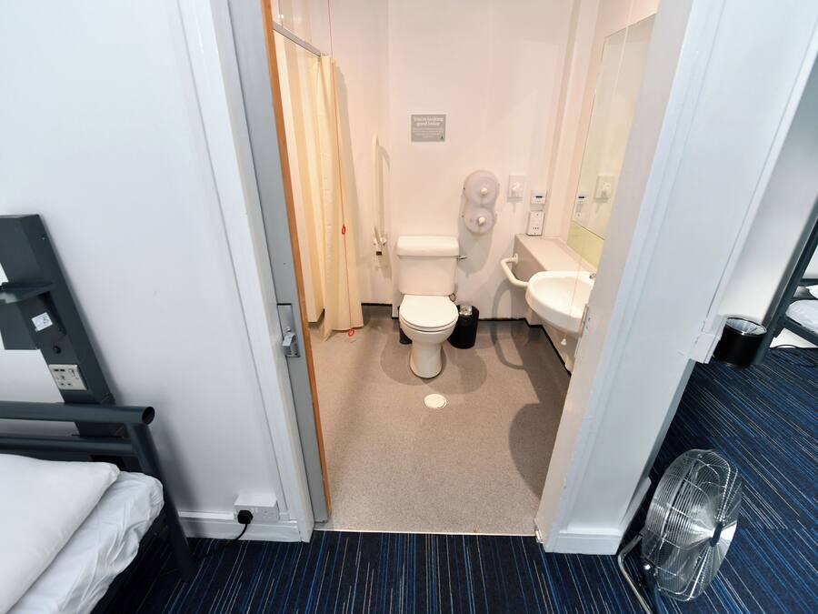 Casa de banho de hotel com sanita, bidé e lavatório. Tem cortina de chuveiro, porta-toalhas e um ventilador.