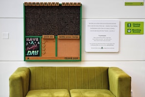 Un tablón de anuncios con un tablón comunitario, una pizarra con mensajes motivadores y un sillón tapizado en verde.