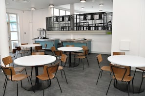 A modern dining area with round tables and chairs, a kitchen area with appliances, and a large window providing natural light. - YHA London Thameside - Hostel (London)