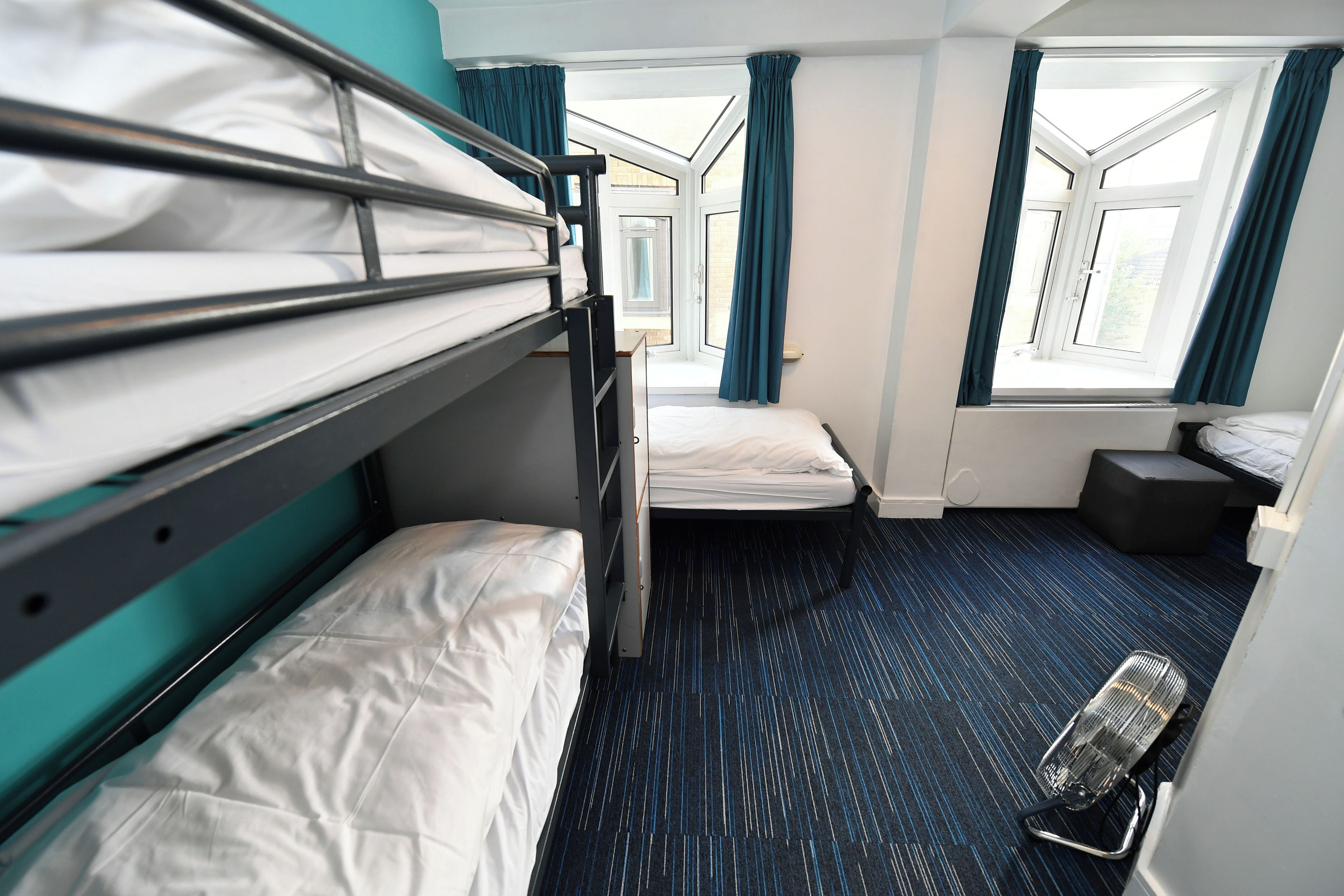 Een kleine hotelkamer met stapelbedden, een klein nachtkastje, een verwarming en ramen met gordijnen.
