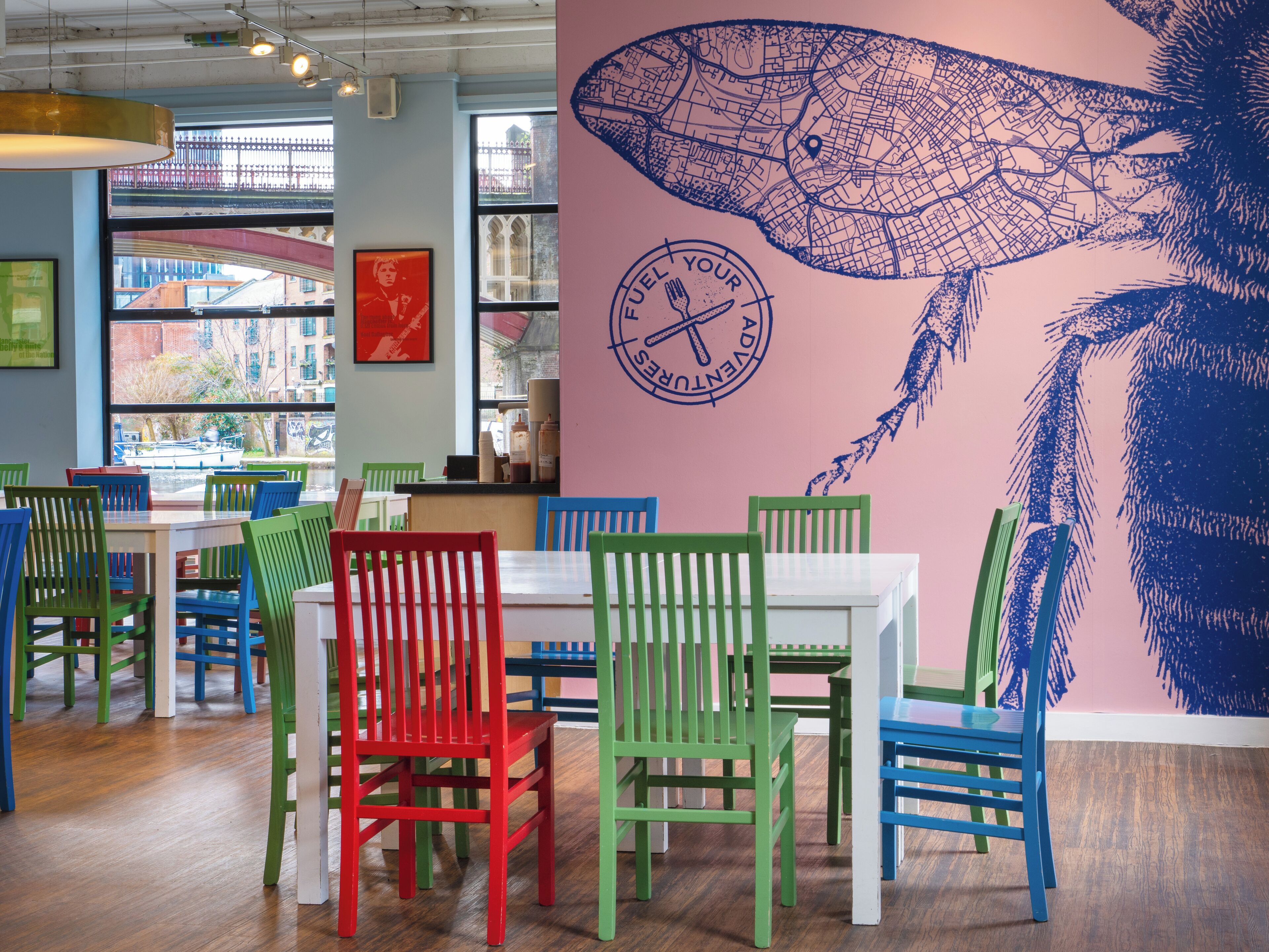 L’intérieur d’un restaurant, avec des chaises aux couleurs vives, une grande fresque murale et une vue sur la rue grâce à de grandes fenêtres.