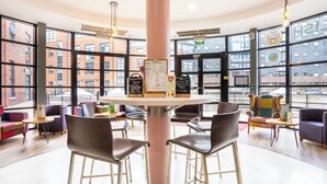 A modern cafe with a central table, bar stools, and large windows offering views of a brick building. - YHA Manchester - Hostel (Manchester)