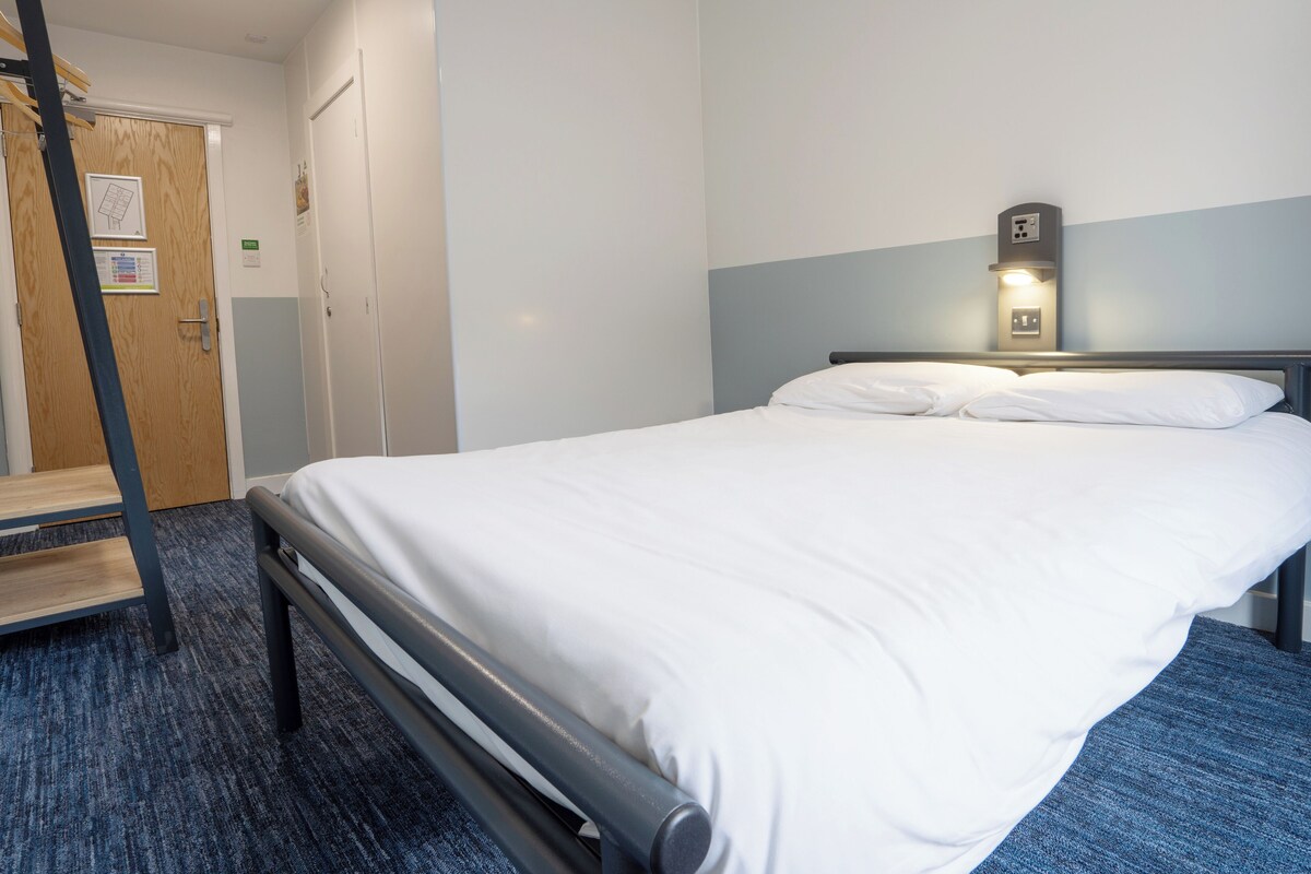 A neatly made bed with white linens, a wooden door with a sign, and a wall-mounted light fixture.