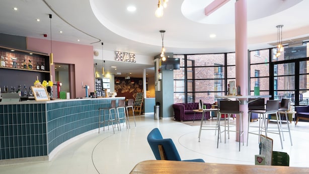 A modern bar area with a curved counter, bar stools, and a seating area with a purple sofa and chairs. - YHA Manchester - Hostel (Manchester)