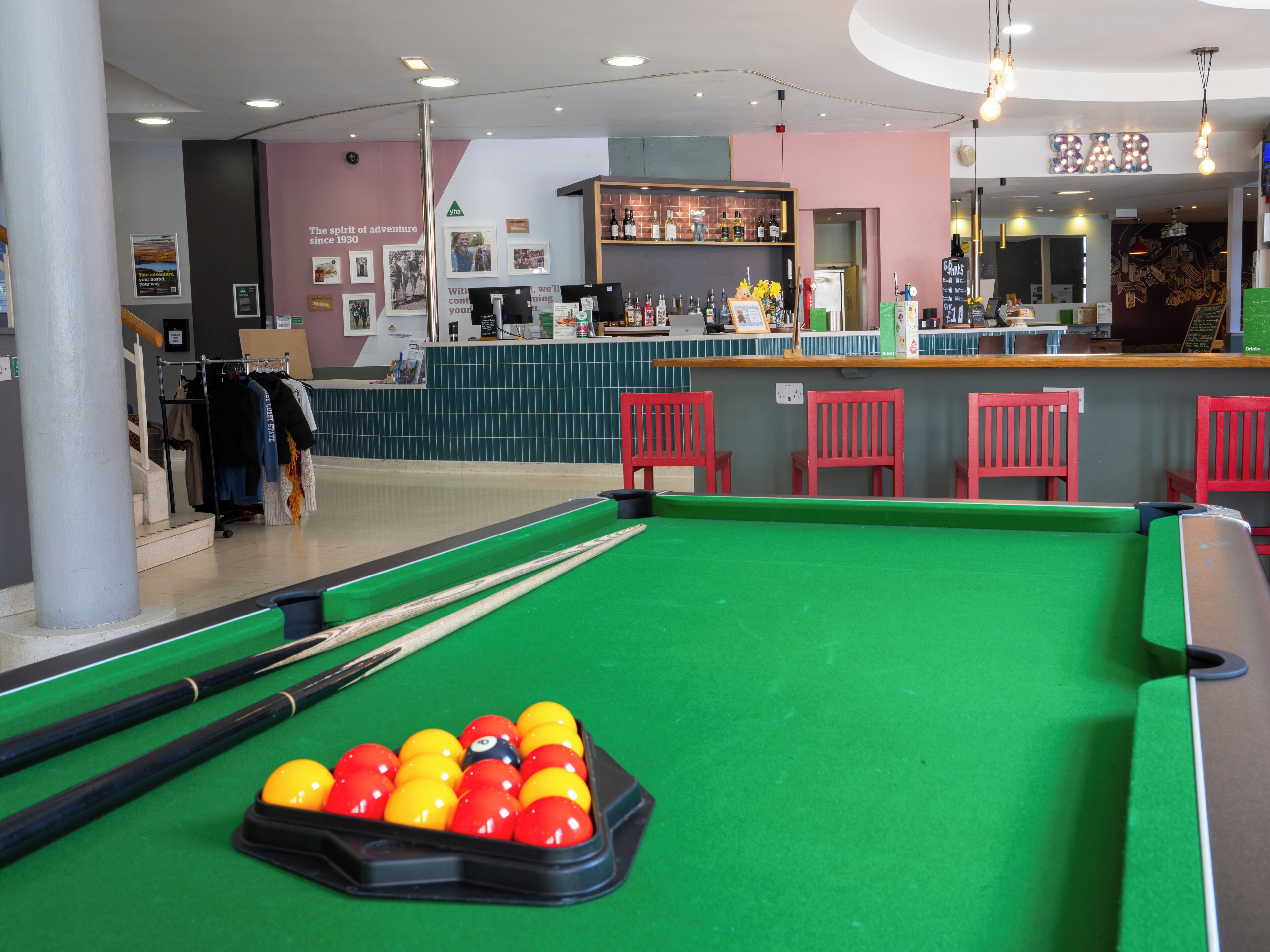 Une table de billard avec des billes, un bar avec une enseigne indiquant « BAR », et un portant à vêtements en arrière-plan.