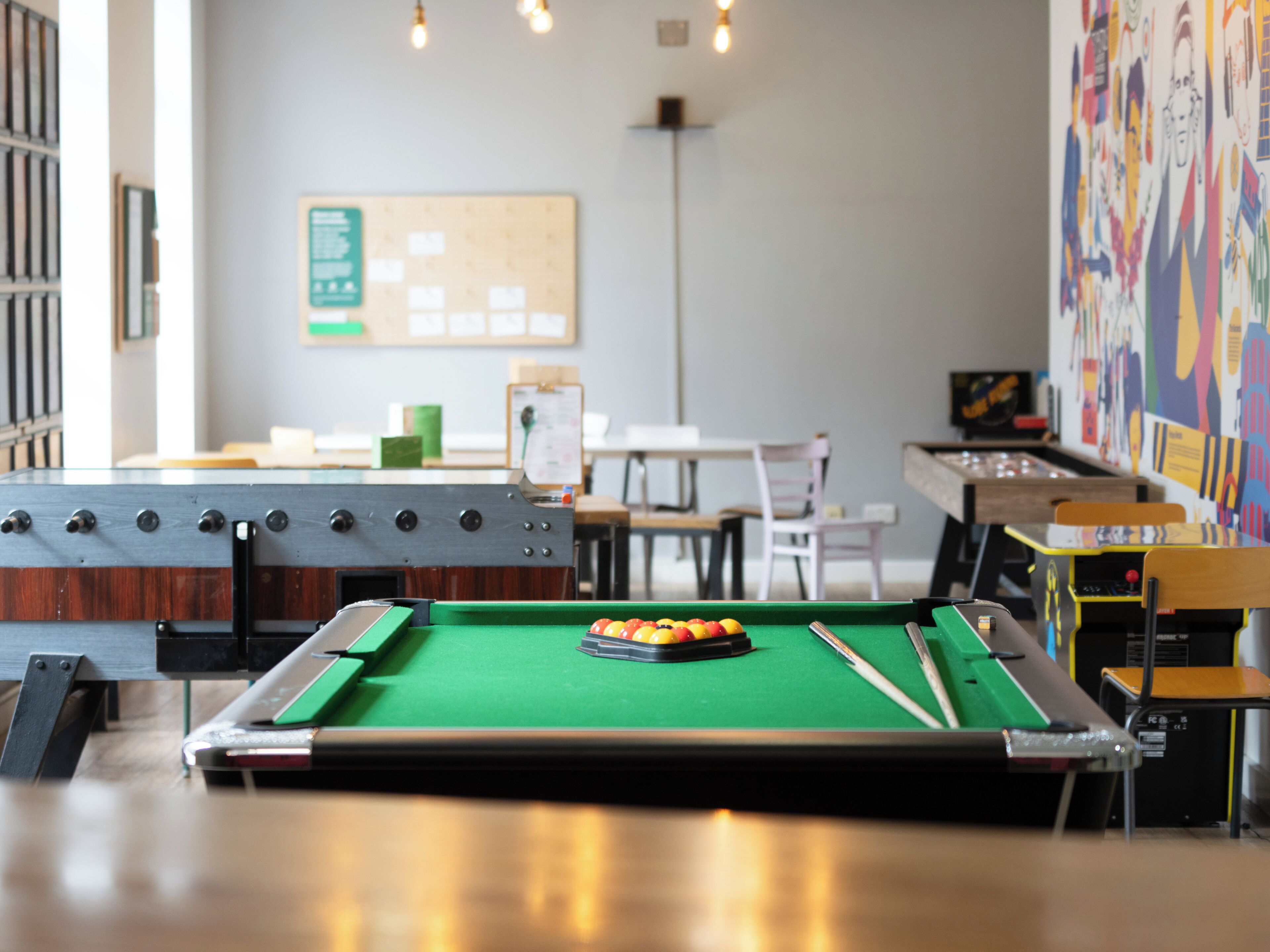 Ein Billardtisch mit Kugeln, ein Kicker und eine Dartscheibe in einem Raum mit Tischen und Stühlen.