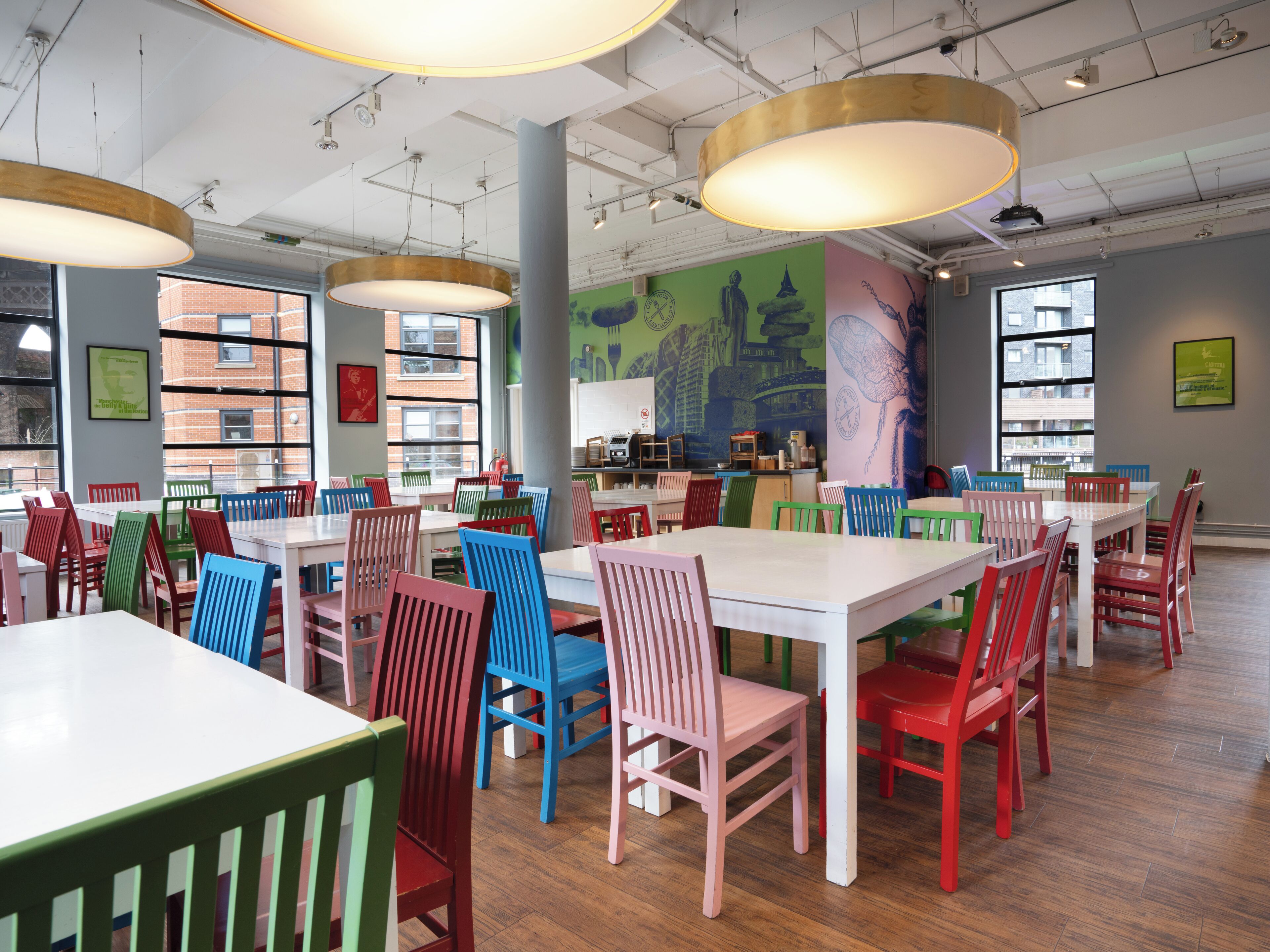 A spacious library with multiple tables and chairs, large windows, and a mural on the wall.