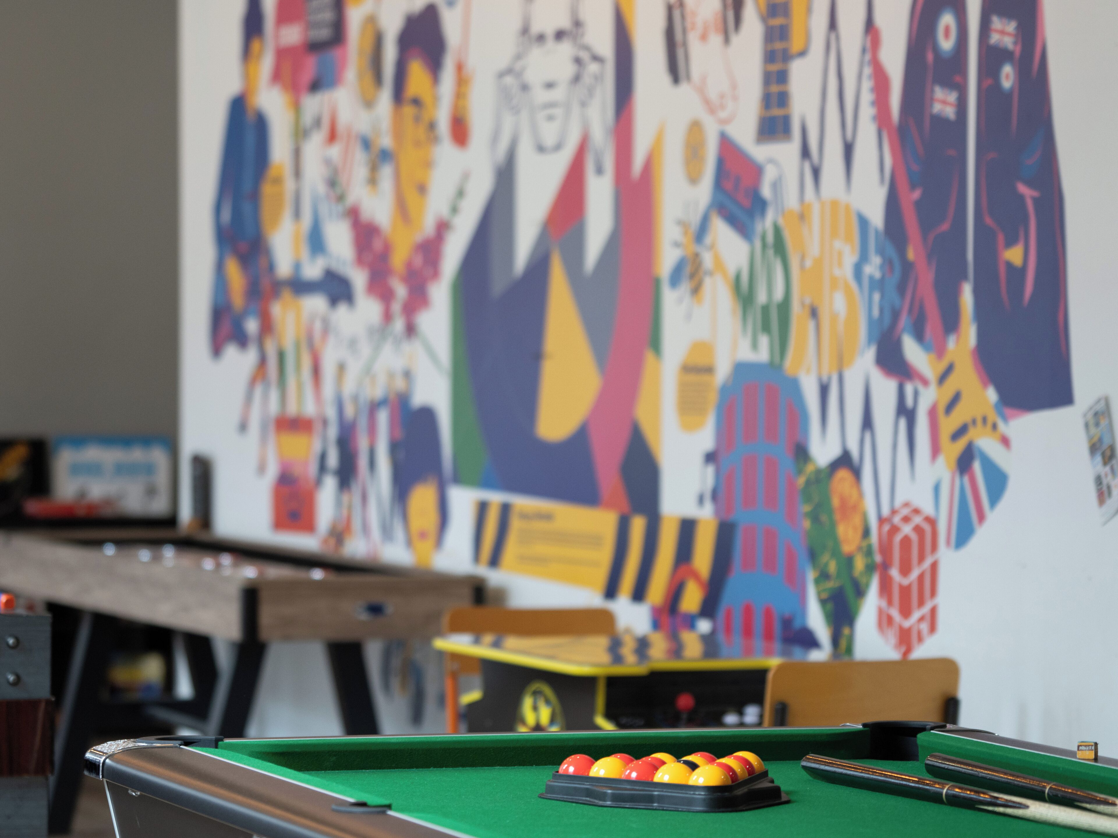 A billiards table with balls and cues, a wooden table in the background, and a colorful abstract mural on the wall.