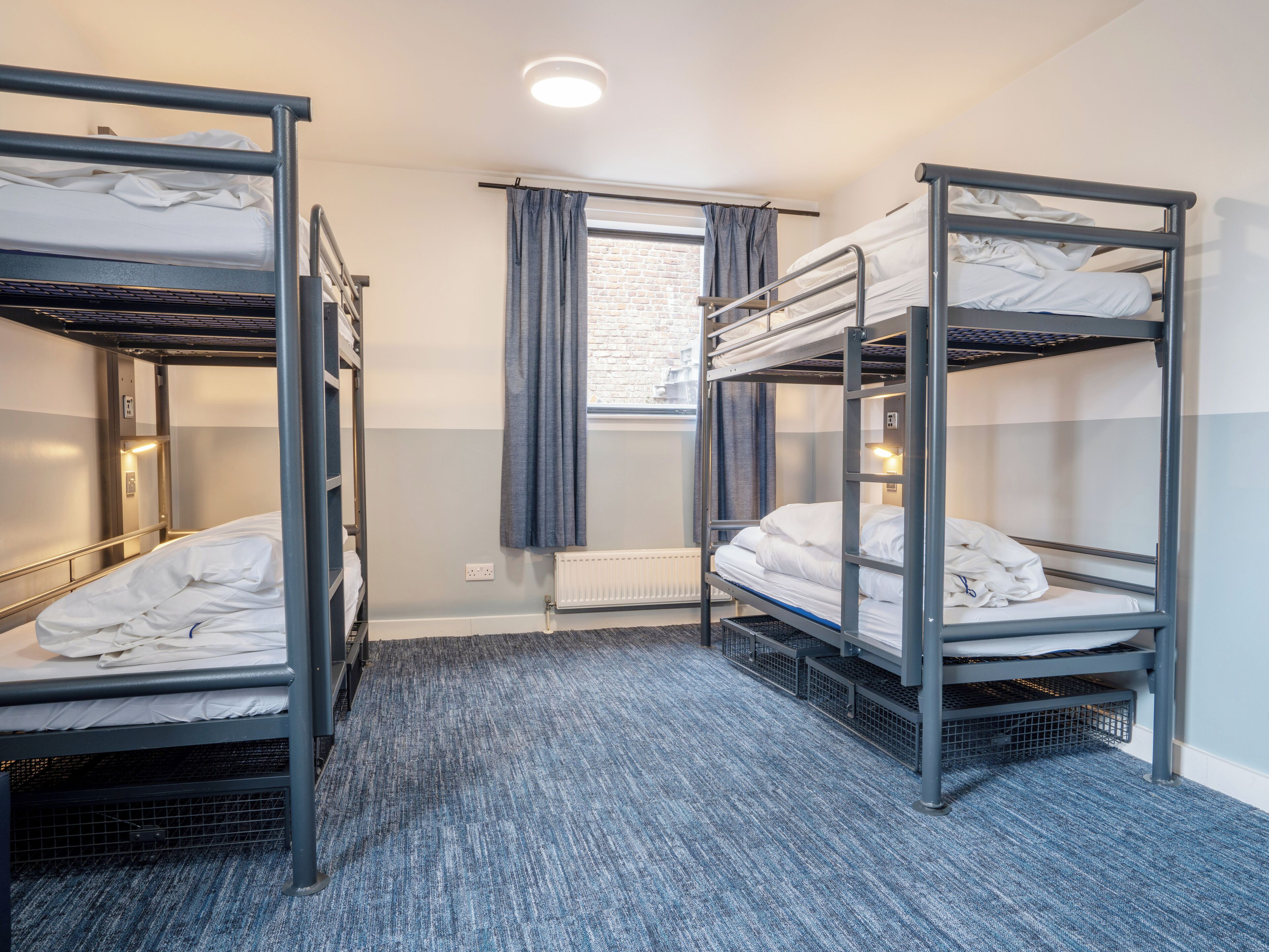 A dormitory room with bunk beds, a window with curtains, a radiator, and a carpeted floor.