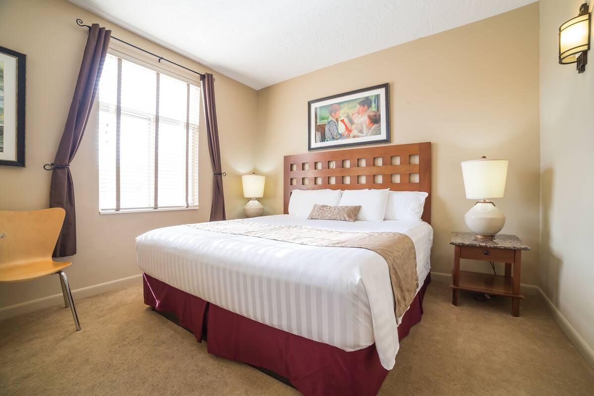 A neatly made bed with a wooden headboard, two bedside lamps, a framed picture on the wall, and a window with curtains.