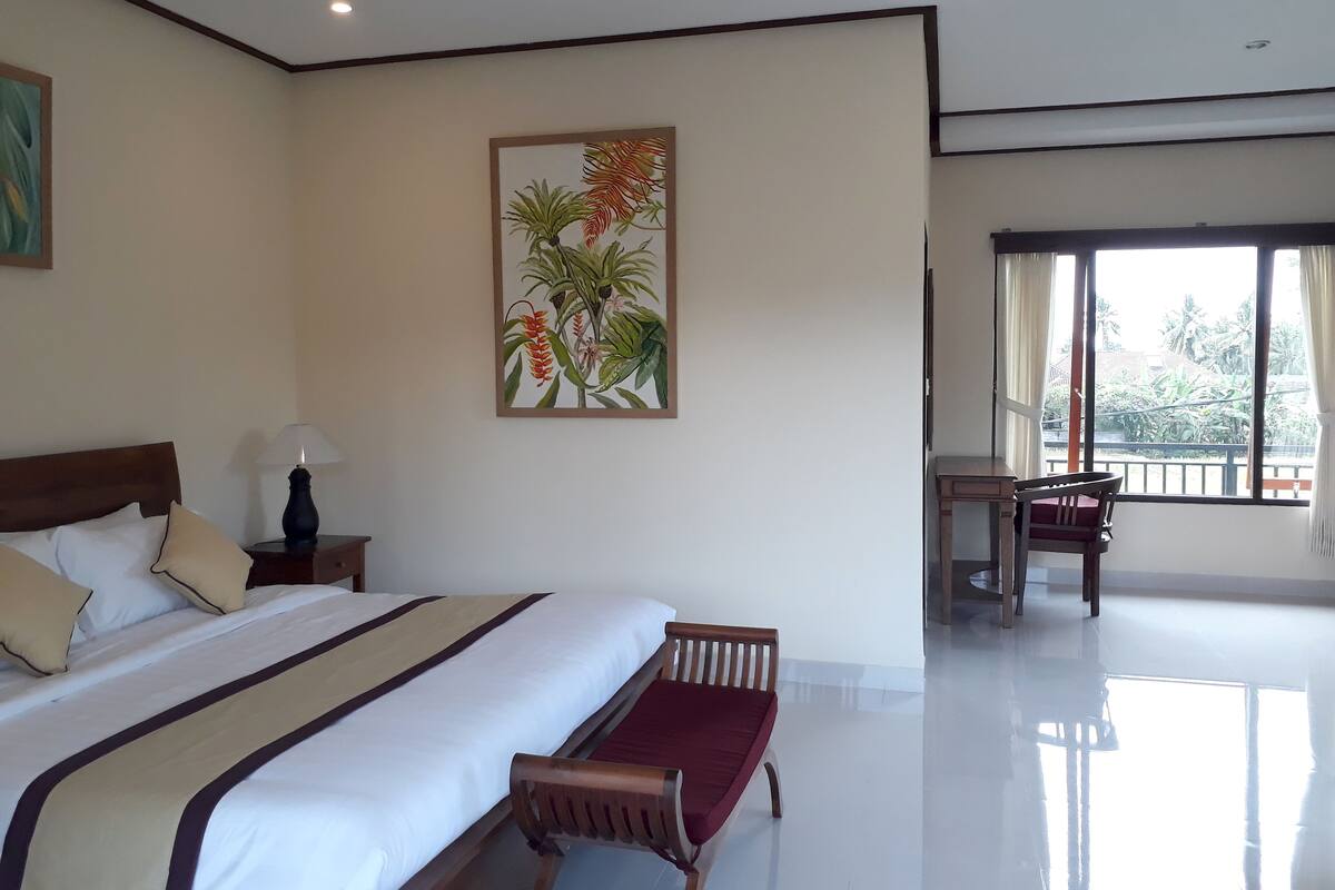 A hotel room with a large bed, a bench, a desk, and a window with a view of palm trees.