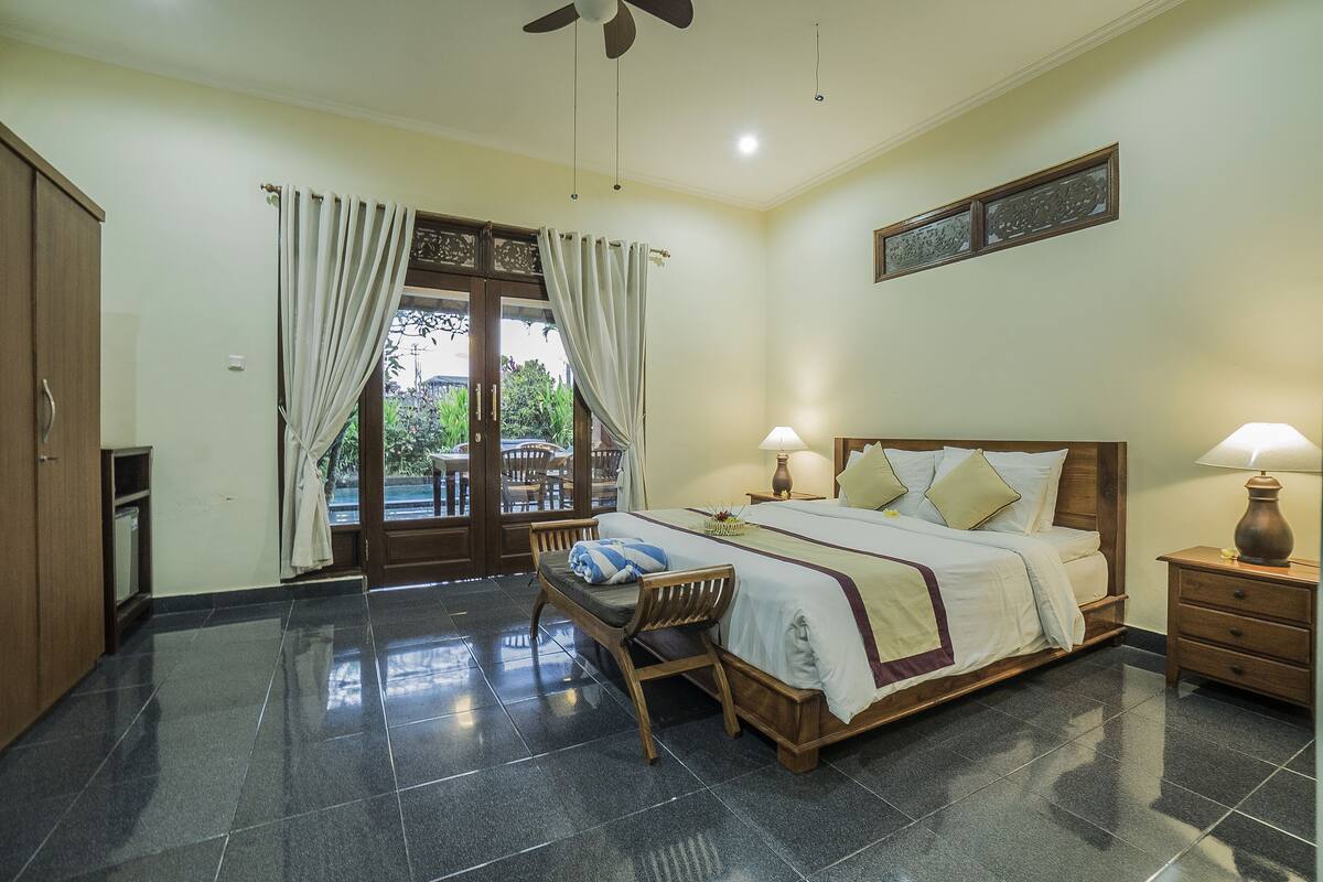 A bedroom with a large bed, two bedside tables with lamps, a wooden wardrobe, a chair, and a view through a glass door.