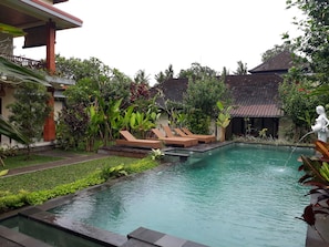 A swimming pool with lounge chairs, surrounded by lush greenery and a traditional building with a tiled roof. - Nyoman Sandi Guest House (Ubud)