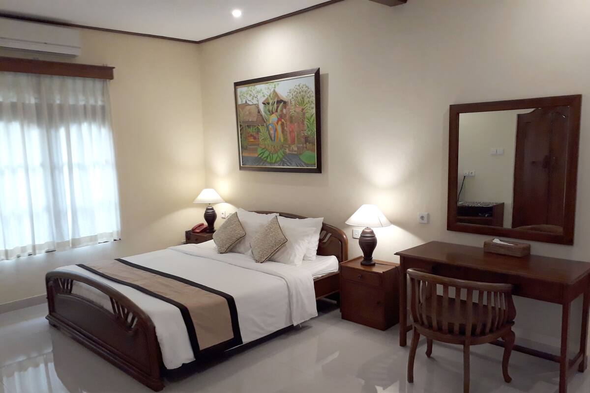 A hotel room with a bed, two bedside tables with lamps, a desk with a chair, a large mirror, and a painting on the wall.