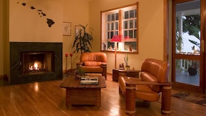 Un salon doté d’une cheminée, de fauteuils en cuir, d’une table basse avec des livres, d’une plante en pot et d’une fenêtre donnant sur un paysage verdoyant.