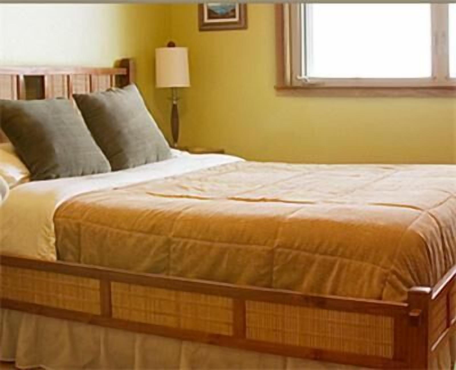 Un lit avec un cadre en bois et une couette beige, une table de chevet avec une lampe, et une fenêtre donnant sur l’extérieur.