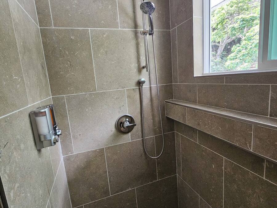 Ducha con cabezal de ducha manual y grifo empotrado en la pared, paredes revestidas de azulejos y ventana con vista a la vegetación.