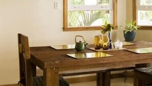 Une table à manger en bois, entourée de chaises, une fenêtre donnant sur un paysage verdoyant et un tableau encadré accroché au mur.