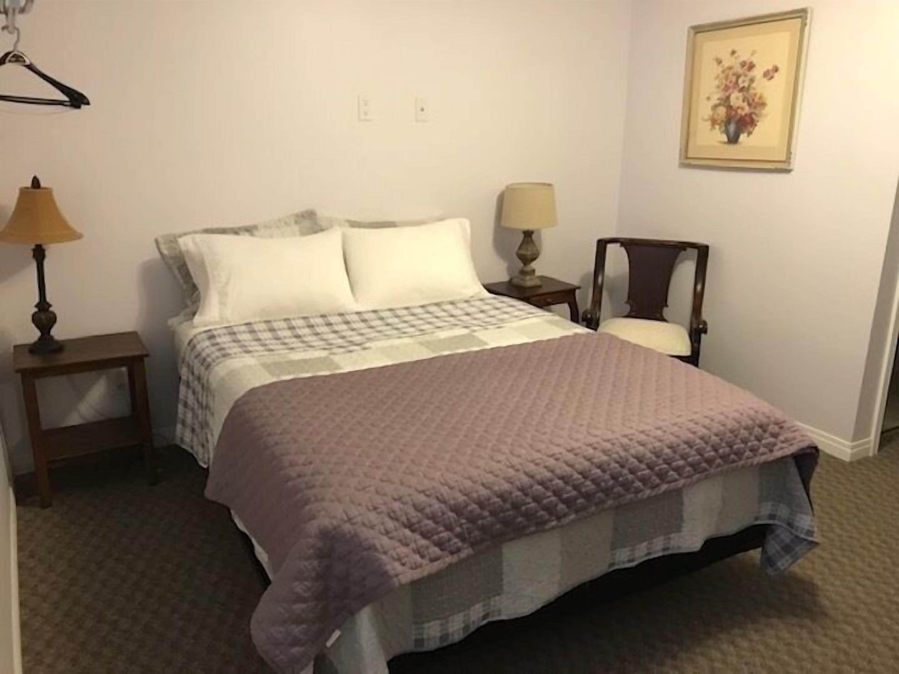 A neatly made bed with a checkered bedspread and a purple quilt, a bedside lamp, a small table, and a chair.