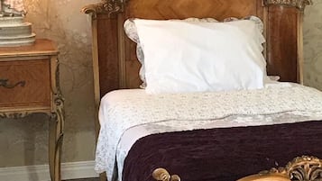 Una cama de madera clásica con un almohadón blanco y una colcha de encaje, sobre un fondo de papel tapiz floral.