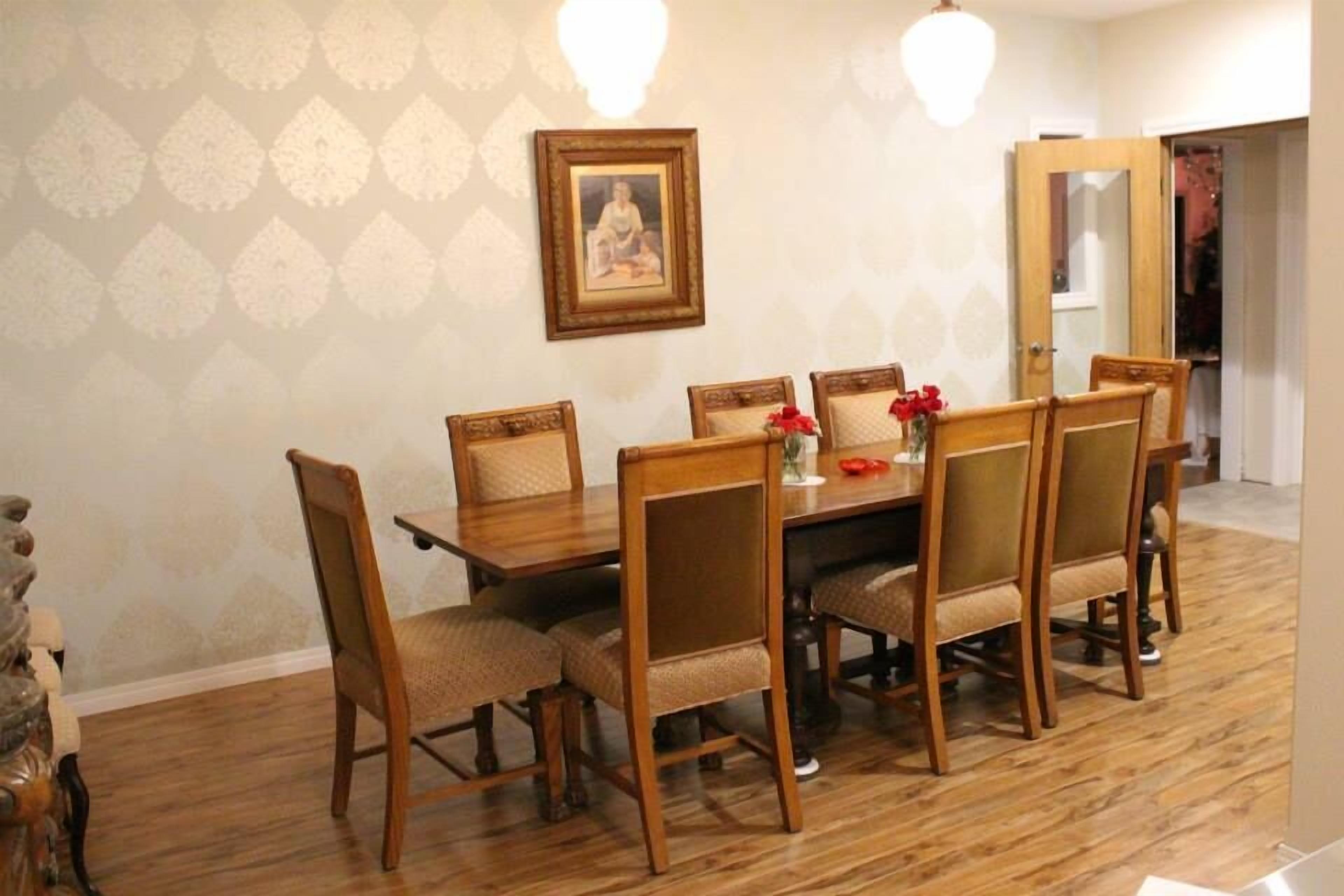A dining area with a wooden table and chairs, a framed picture on the wall, and a vase with flowers on the table.