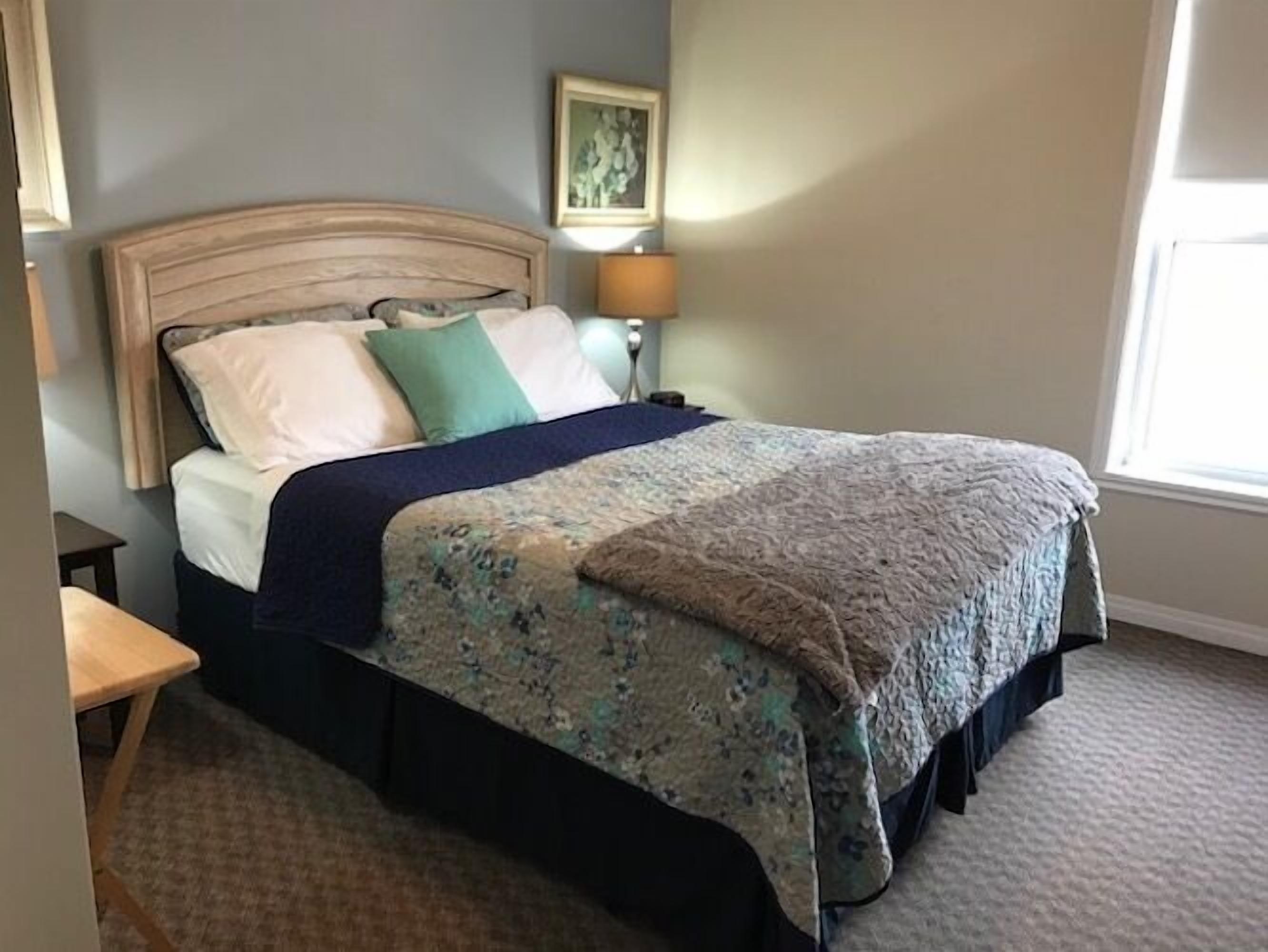 Een net opgemaakt bed met een bedovertrek met patroon en kussens, een bedlampje en een ingelijst beeld aan de muur.
