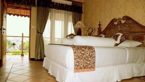 A double bed with a patterned throw, a nightstand with a lamp, and a view of a balcony with a railing. - Omah Sinten Heritage Hotel & Resto (Surakarta)