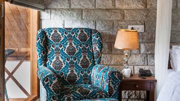 A room with a patterned armchair and ottoman, a wooden ceiling, stone wall, and a nightstand with a lamp and phone.
