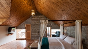 A bedroom with a wooden ceiling, a brick wall, a bed with white linens, a green throw blanket, and a television on a shelf. - Larisa Resort, Manali (Manali)