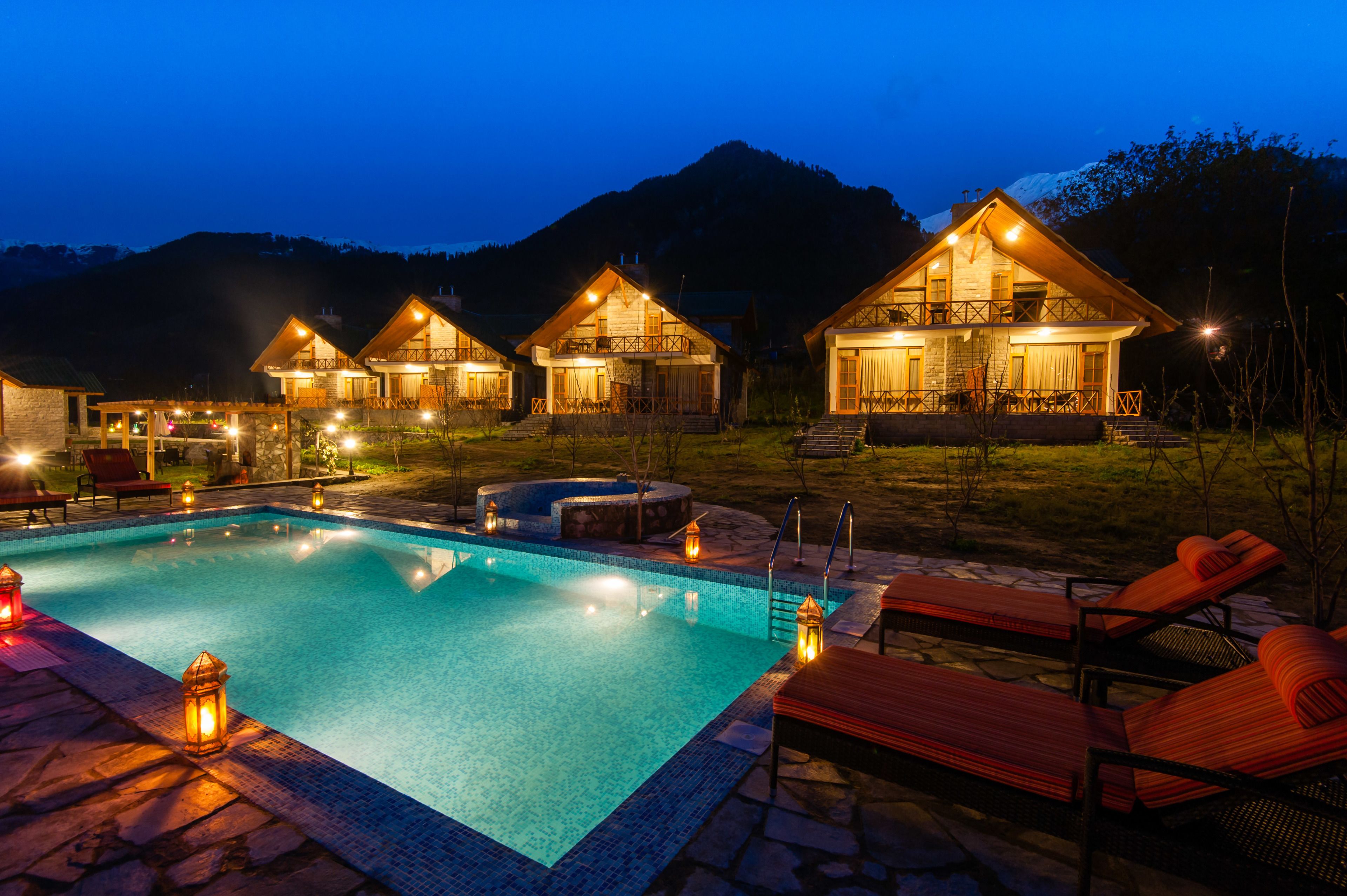 A pool area with lounge chairs and a hot tub, surrounded by wooden cabins with balconies, set against a mountainous backdrop.