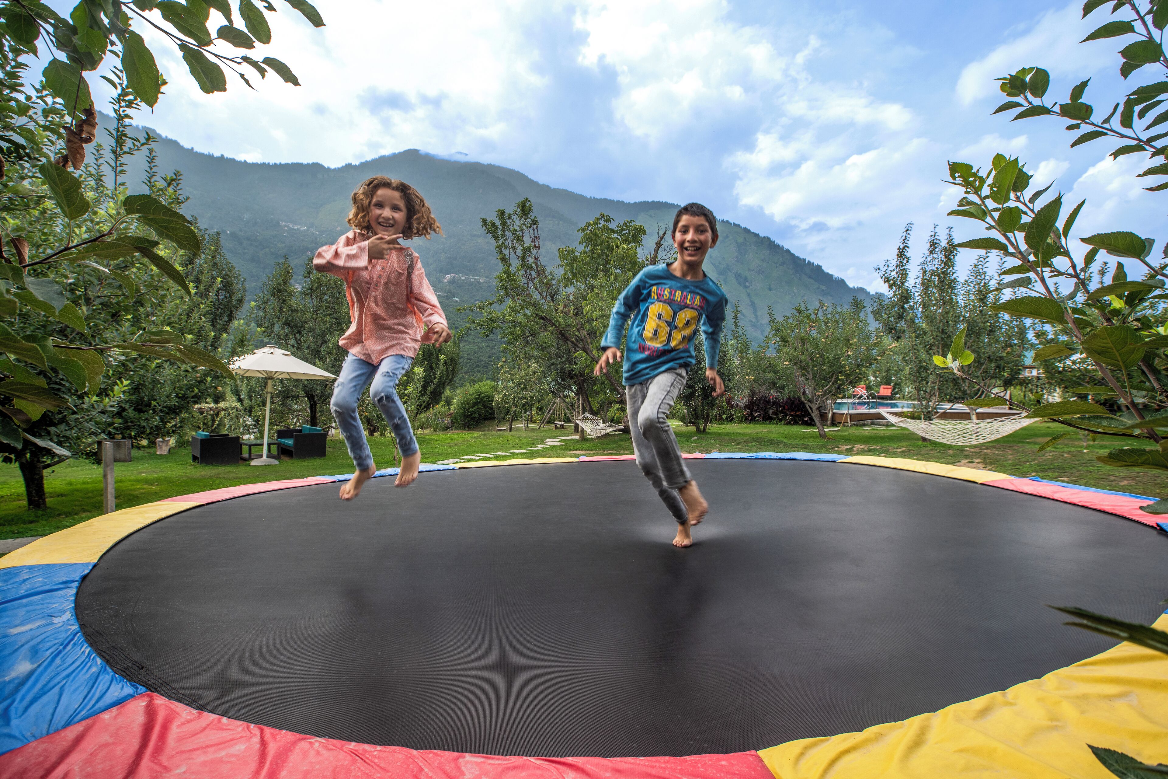 Dos niños saltando en un trampolín rodeado de vegetación y montañas.