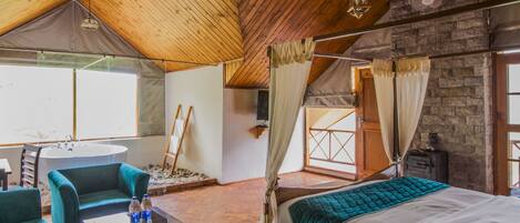 A cozy bedroom with a wooden ceiling, a large bed, a sitting area with chairs, and a view of the outside.