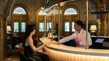 A bar area with a curved counter, a bartender, and a customer. There are bottles and glasses on the counter, and a cash register is visible.