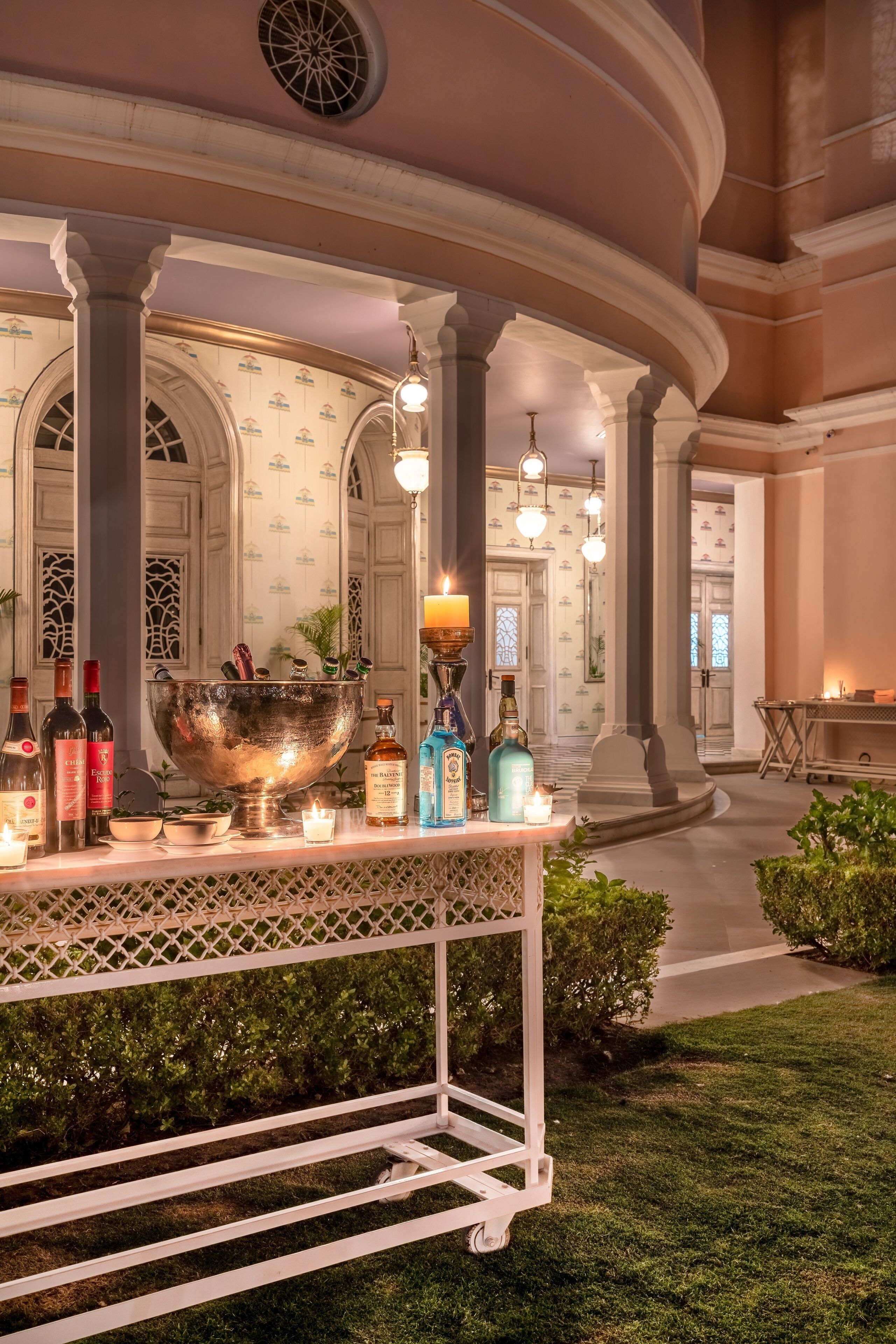 Zona de bar al aire libre bien iluminada con un recipiente metálico grande como centro de atención, rodeada de botellas de licor, velas y una mesa con celosía blanca.