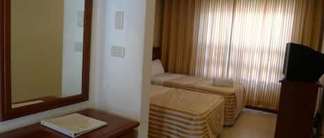 A hotel room with a wooden desk, a mirror, two beds with striped bedding, a television on a stand, and a window with curtains.