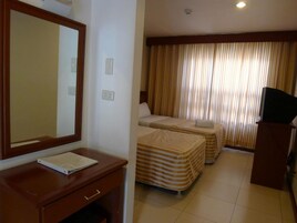A hotel room with a wooden desk, a mirror, two beds with striped bedding, a television on a stand, and a window with curtains.