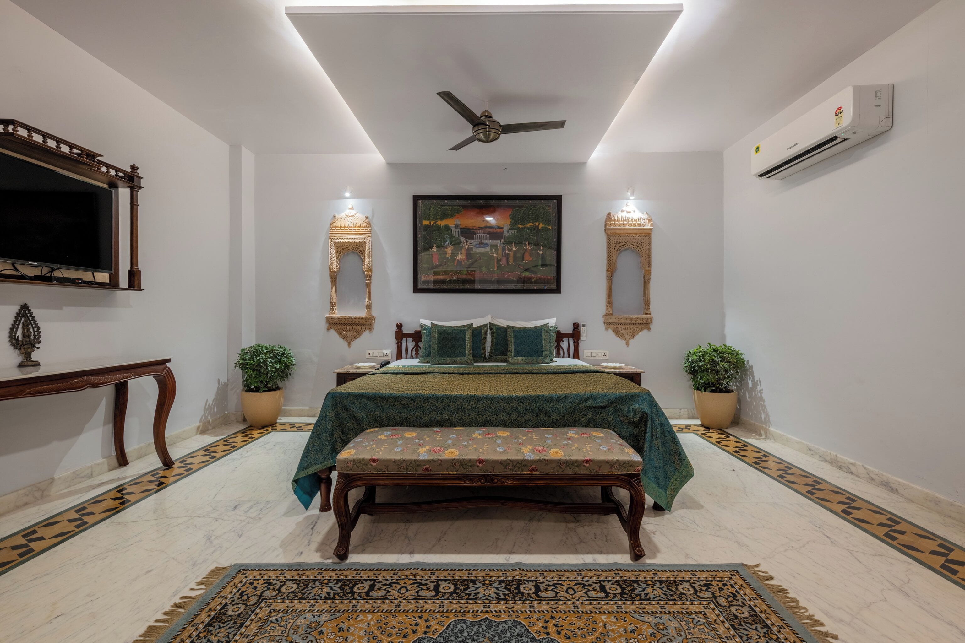A bedroom with a bed, bench, decorative pillows, wall-mounted lights, a ceiling fan, and a framed painting.