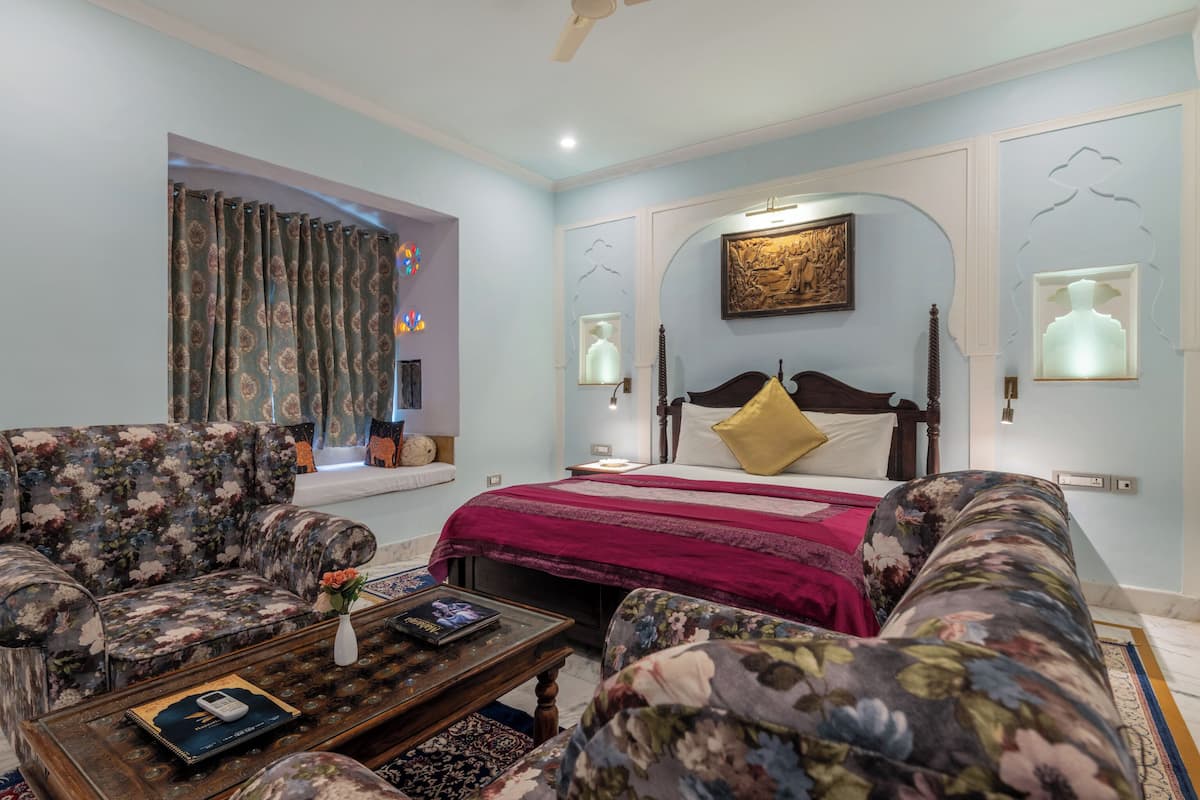 A bedroom with a bed, two floral armchairs, a wooden coffee table, and a window with curtains.
