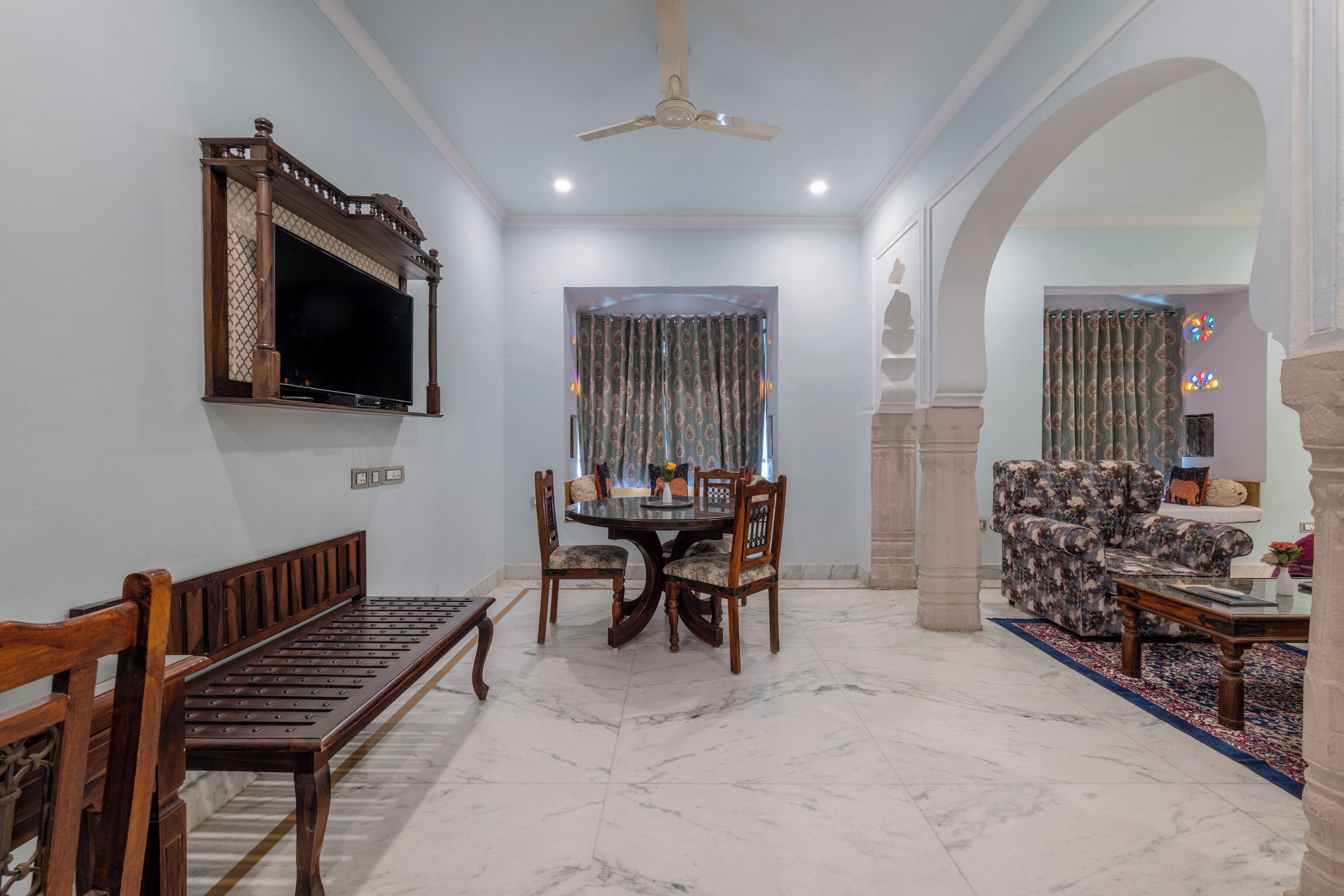 A spacious living area with a marble floor, a dining table with chairs, a sofa, a television mounted on the wall, and a ceiling fan.