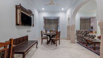 A spacious living area with a marble floor, a dining table with chairs, a sofa, a television mounted on the wall, and a ceiling fan.