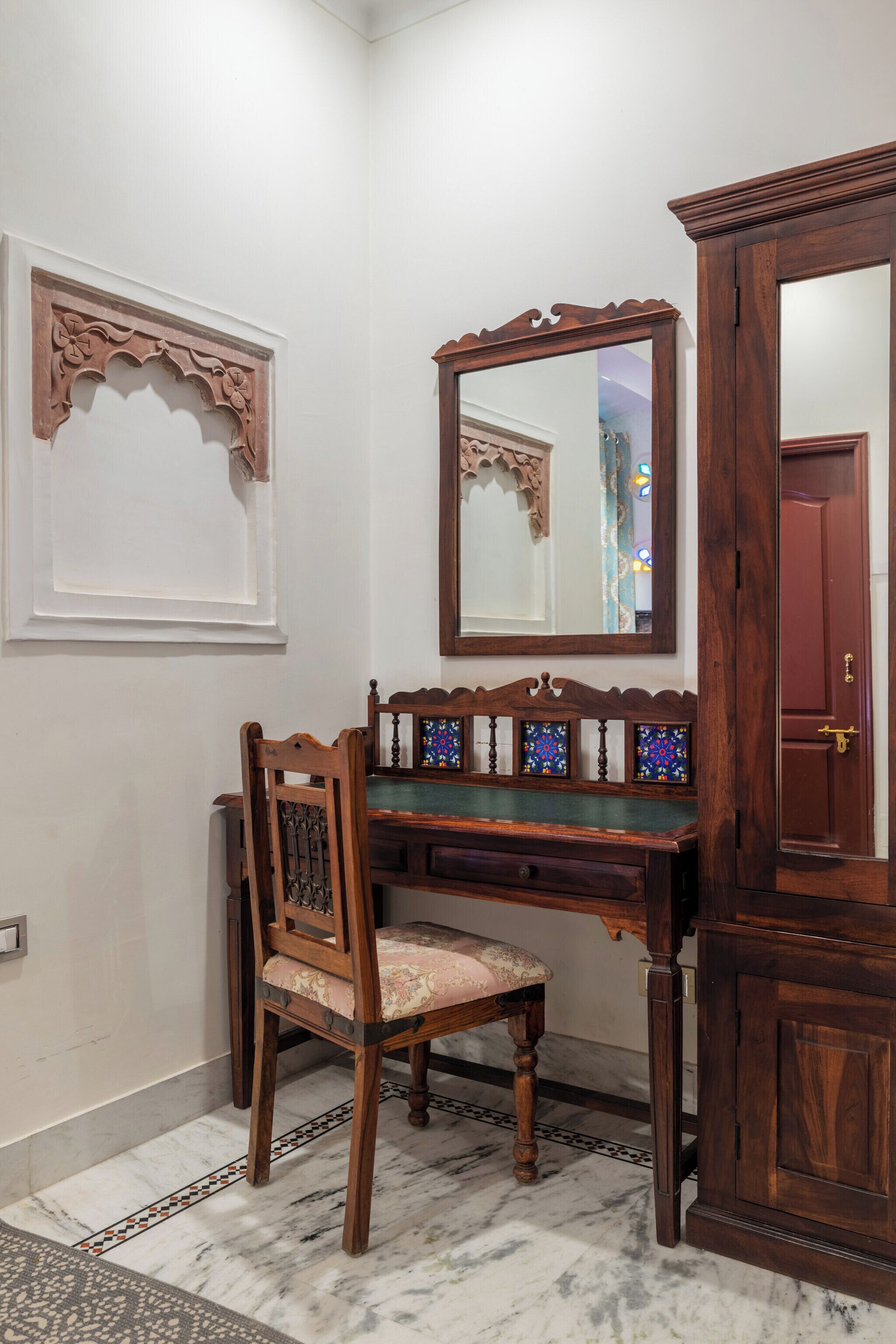 A wooden desk with a chair, a mirror, and a cabinet in a room with white walls and a marble floor.