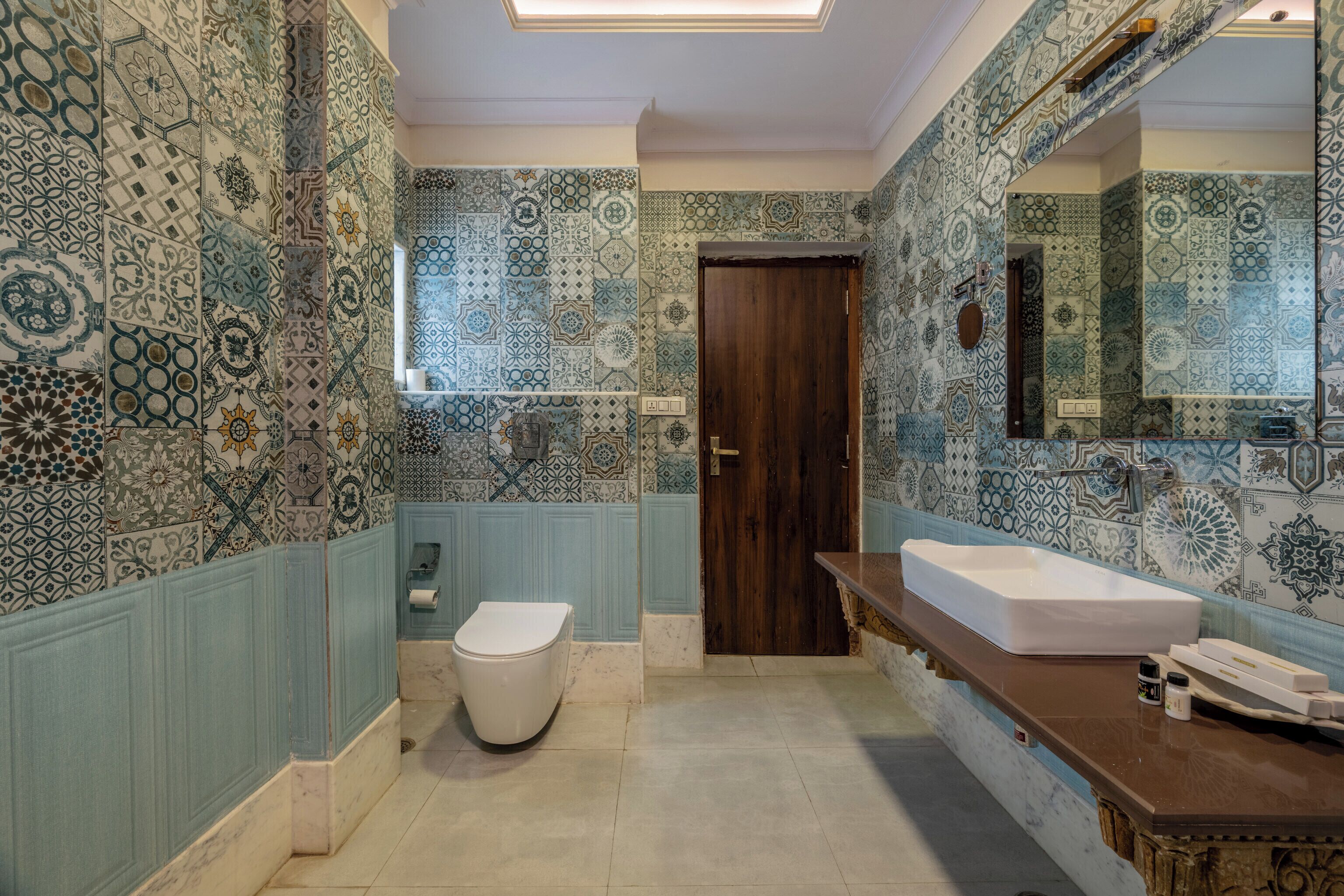 A bathroom with patterned tiles, a wooden door, a white toilet, and a large mirror.